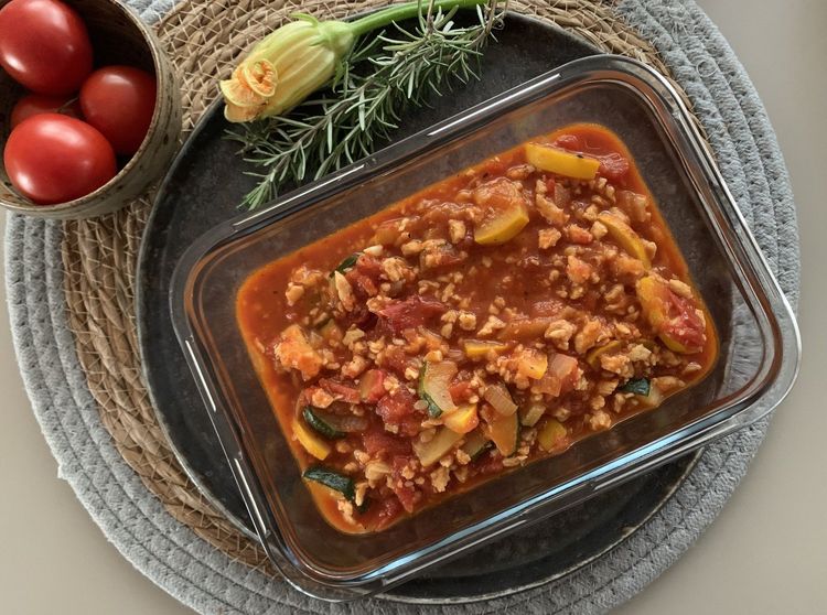 Glasbehälter mit Tomatengemüse und Tofu auf Tablett neben Tomatenkorb und Kräutern