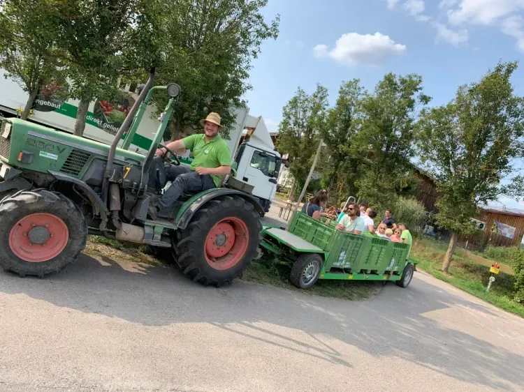 Mann auf grünem Traktor zieht Anhänger mit mehreren Fahrgästen bei sonnigem Wetter auf Landstraße