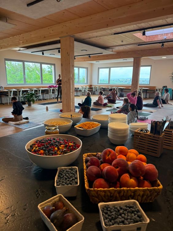Yoga-Gruppe in hellem Raum mit großen Fenstern, Obstschalen und Schüsseln mit Müsli im Vordergrund