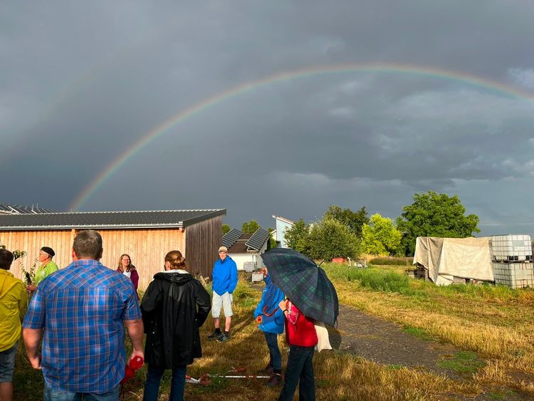 Menschen schauen auf einen Regenbogen über einer ländlichen Szene mit Gebäuden und Bäumen bei Regenwetter