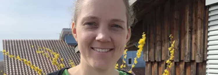 Lächelnde Frau im Grünen vor Bauernhaus und blühendem Strauch an sonnigem Tag