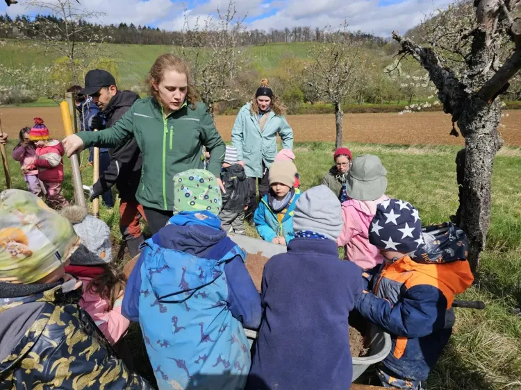 Kinder und Erwachsene beim gemeinsamen Pflanzen im Frühling auf einer Wiese mit blühenden Bäumen