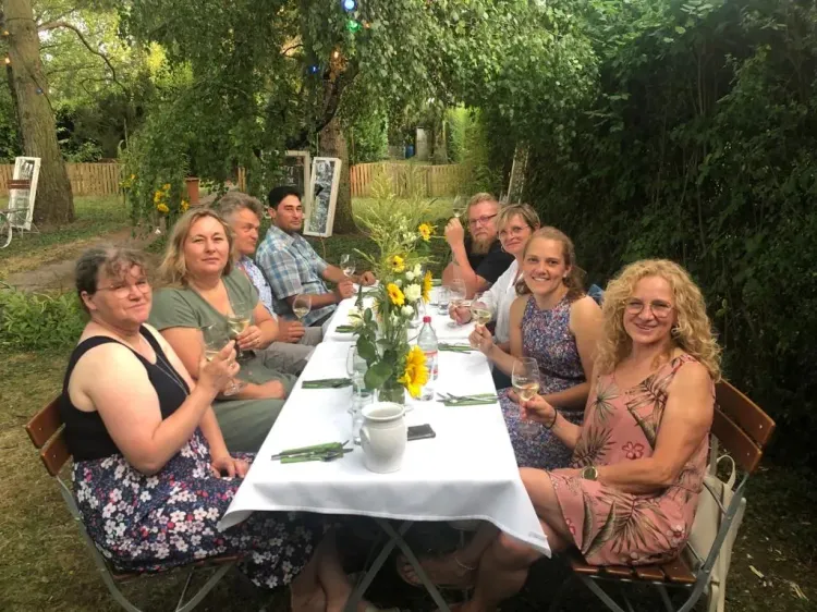 Gruppenfoto von acht Menschen, die an einem Tisch im Freien mit weißen Tischdecke sitzen und Wein trinken.