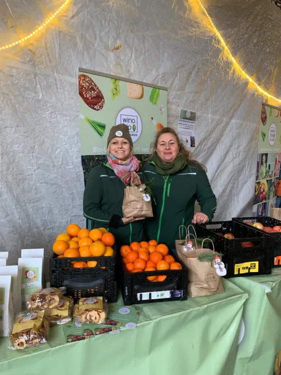 Zwei Frauen an einem Marktstand mit Orangen, Äpfeln und Bio-Produkten in Papiertüten
