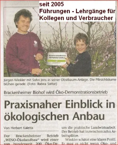 Jürgen Winkler und Sohn Jens zeigen Schild vor blühenden Pfirsichbäumen in Obstbaum-Anlage