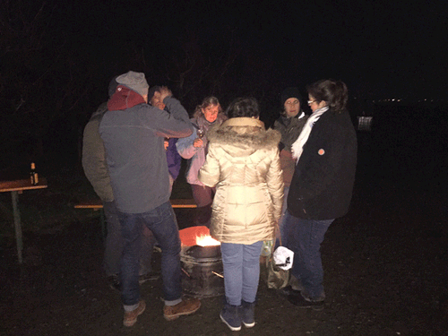 Gruppe von Menschen steht nachts um ein Lagerfeuer im Freien, trägt warme Winterkleidung