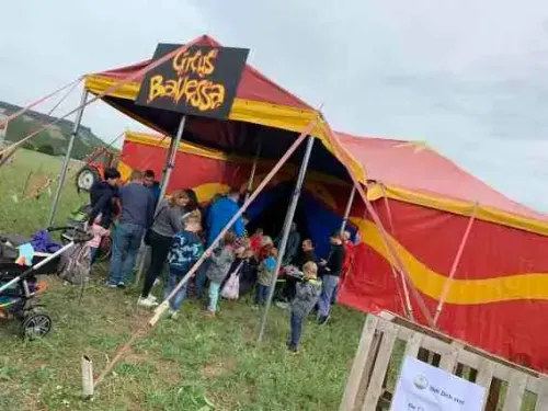 Kinder und Erwachsene warten vor dem Zirkuszelt mit dem Schild Zirkus Balessa auf einer Wiese.