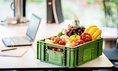 Grüne Kiste mit frischem Obst und Gemüse auf weißem Tisch neben Laptop.